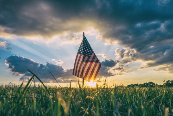 An American flag atop a pole in a green grassy field with the sun rising behind it.